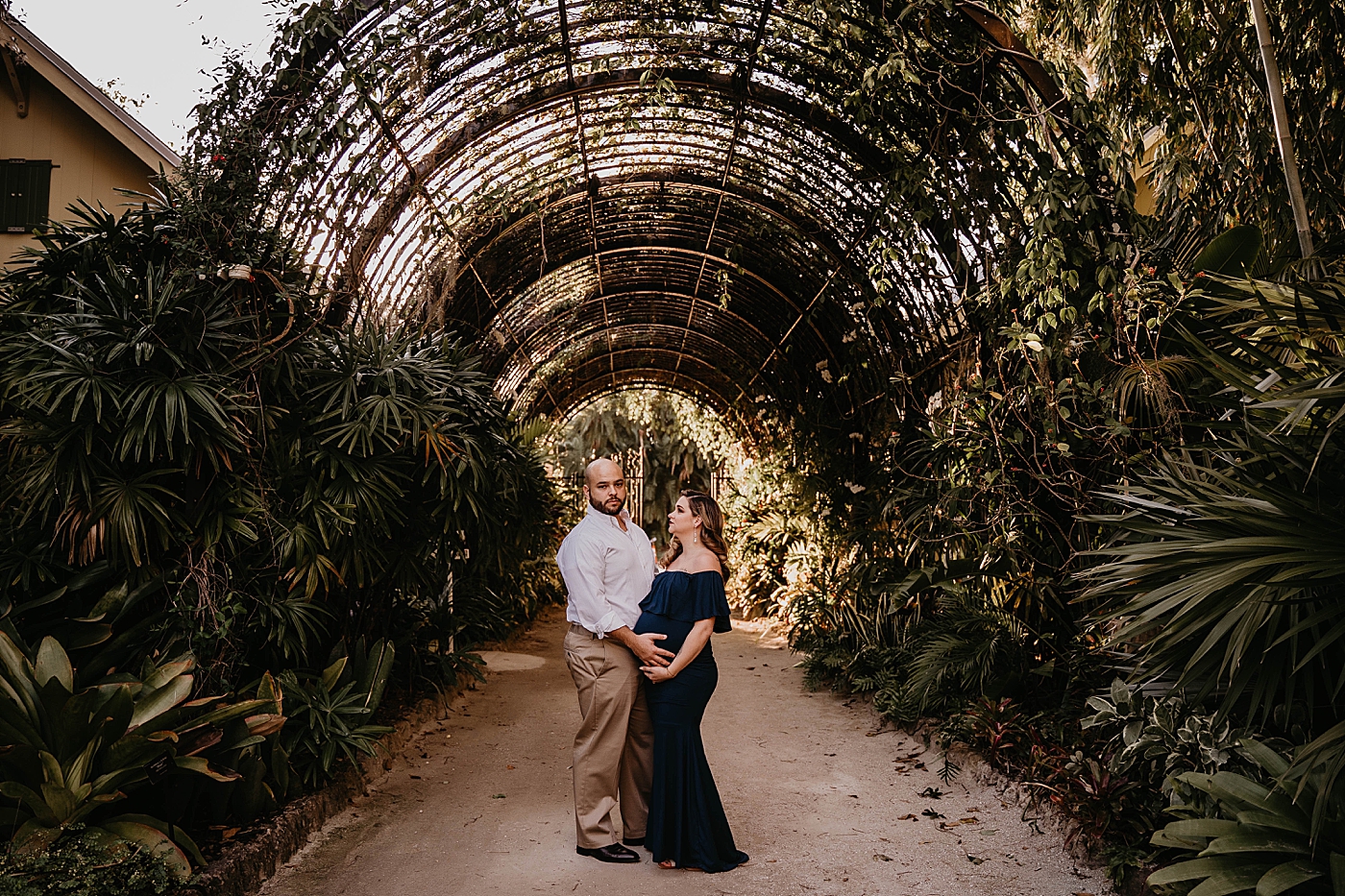 Husband and wife holding bump under green arch way South Florida Maternity Photography captured by South Florida Family Photographer Krystal Capone Photography 