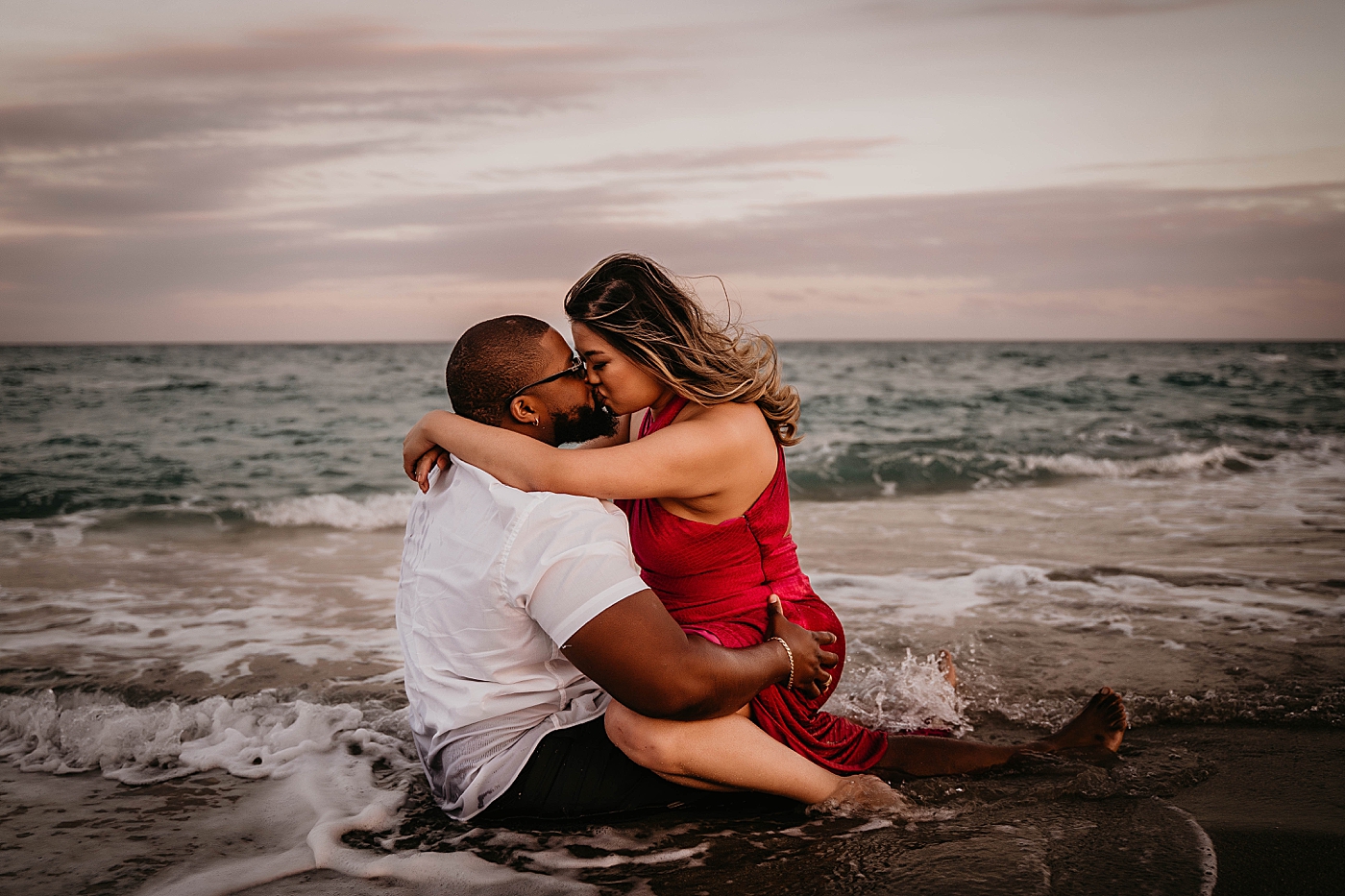 Wife sitting on husband's lap as waves come in and they kiss Palm Beach Anniversary Photography captured by South Florida Family Photographer Krystal Capone Photography
