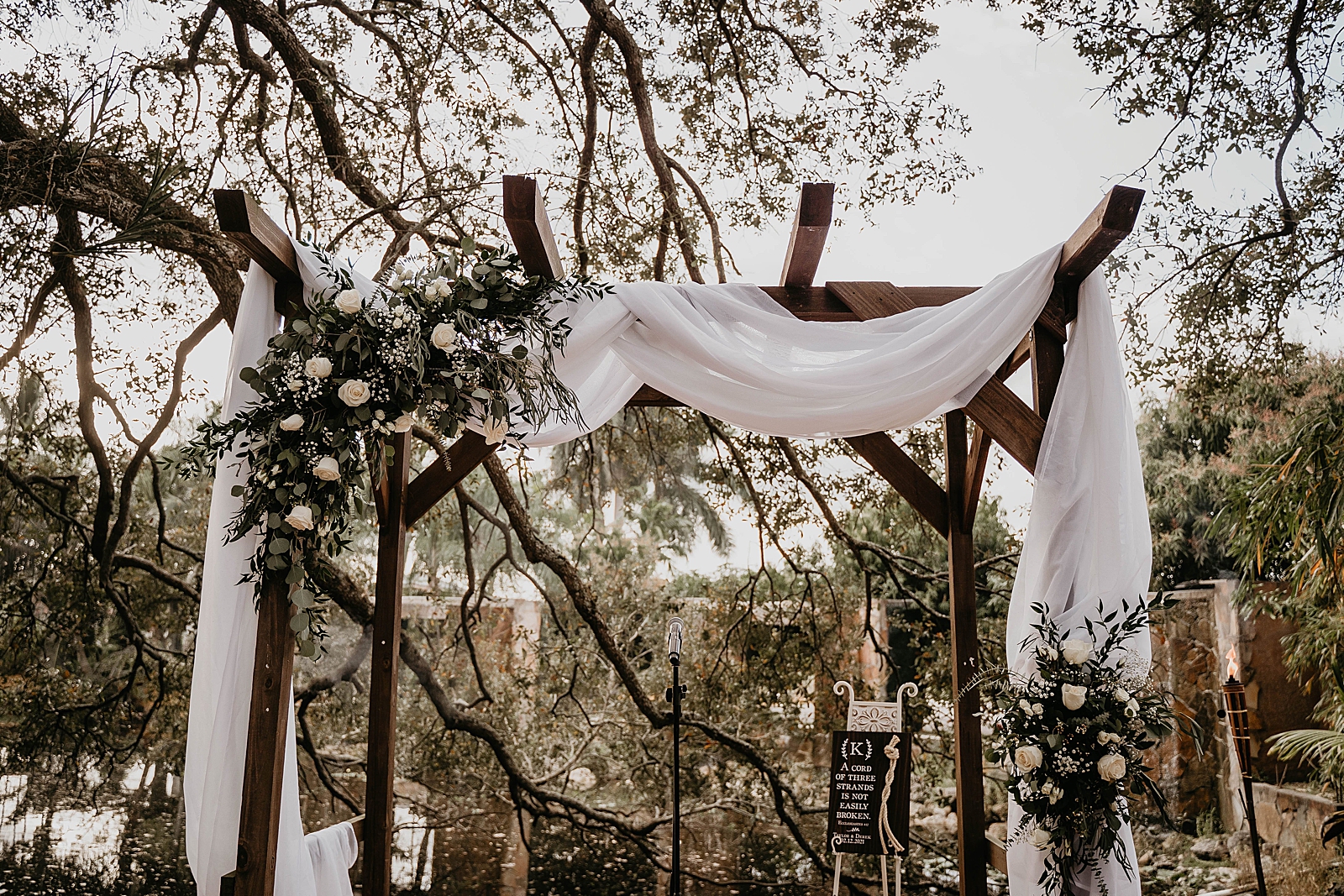 Detail shot of wooden archway with white veil and white flowers Living Sculptures Sanctuary Wedding Photography captured by South Florida Wedding Photographer Krystal Capone Photography