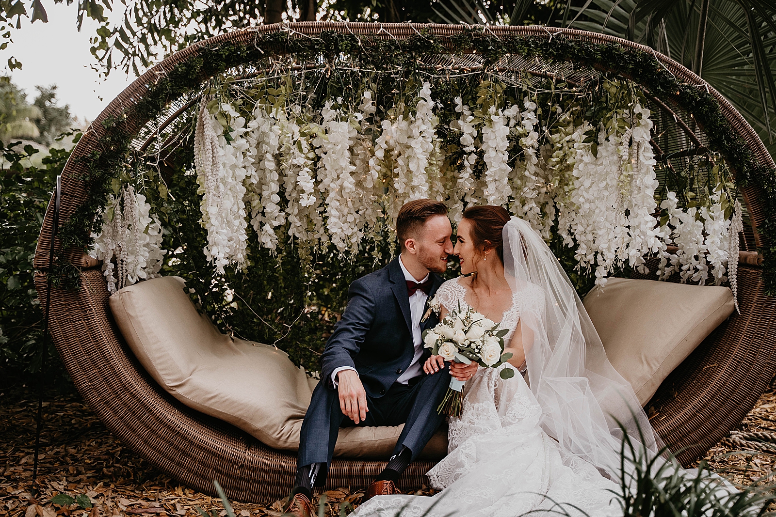 Couple sitting on Garden couch nuzzling noses with white petals hanging Living Sculptures Sanctuary Wedding Photography captured by South Florida Wedding Photographer Krystal Capone Photography