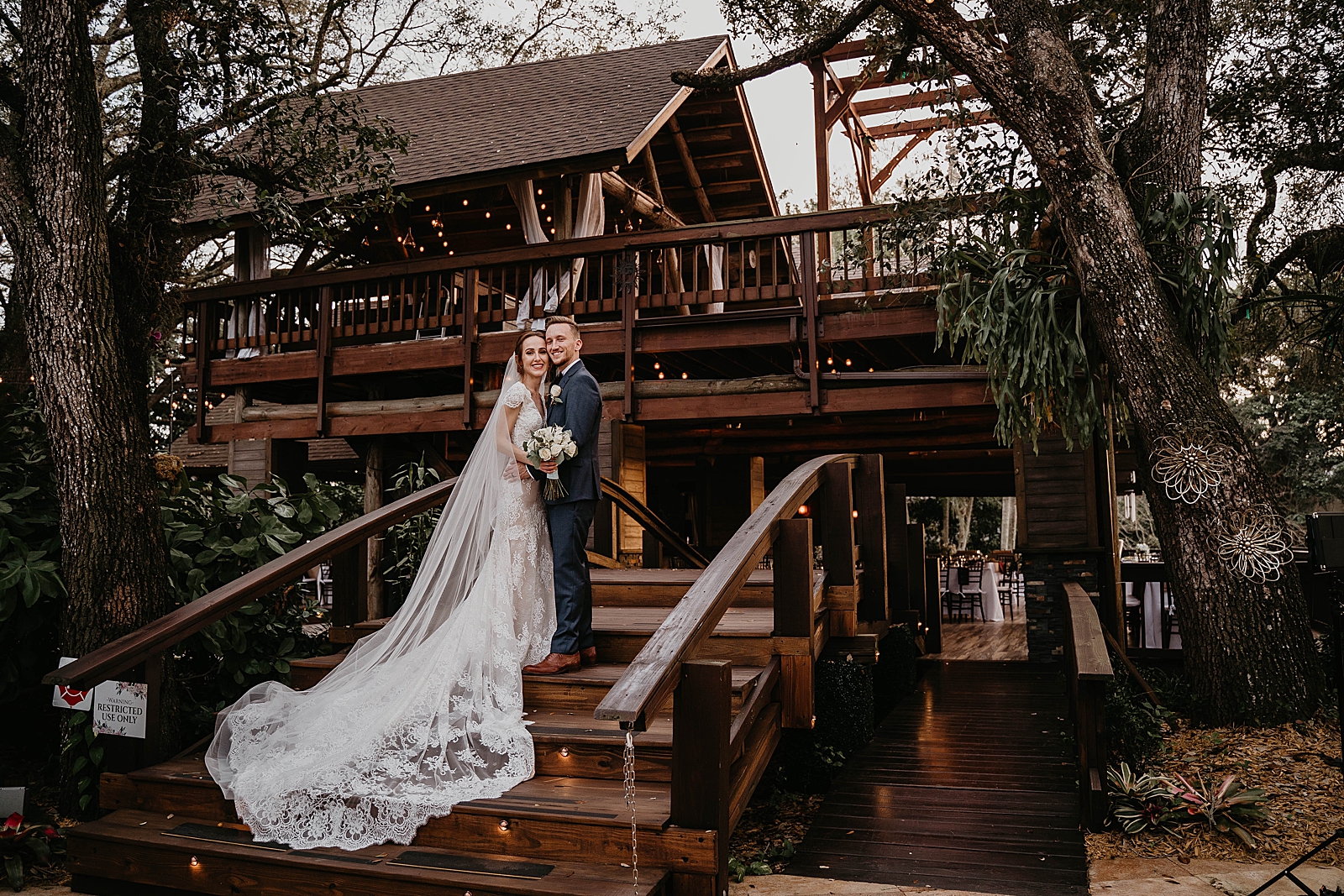 Bride and Groom holding each other close on wooden stairs to large wood lodge with veil flowing down Living Sculptures Sanctuary Wedding Photography captured by South Florida Wedding Photographer Krystal Capone Photography