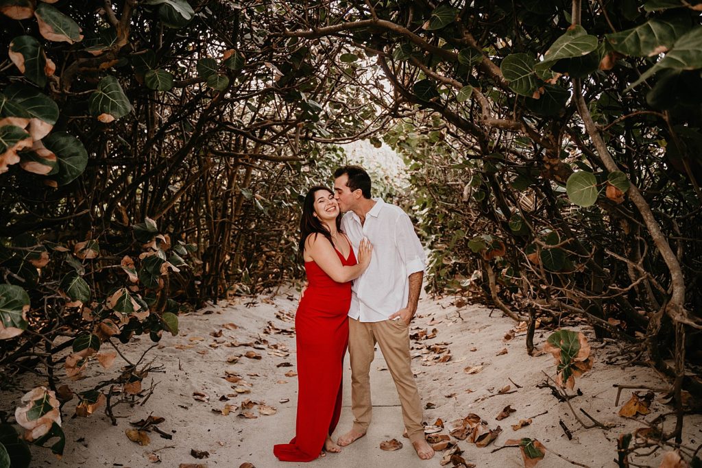 Man kisses woman on the cheek as they stand on sand surrounded by branches with green leafs Palm Beach Engagement Photography captured by South Florida Engagement Photographer Krystal Capone Photography 