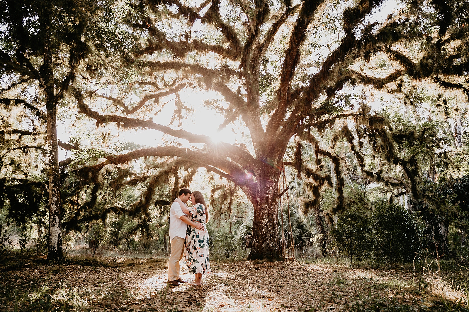 Couple holding each other close with big old spanish moss tree with sun beams shining through Riverbend Park Engagement Photography captured by South Florida Engagement Photographer Krystal Capone Photography 