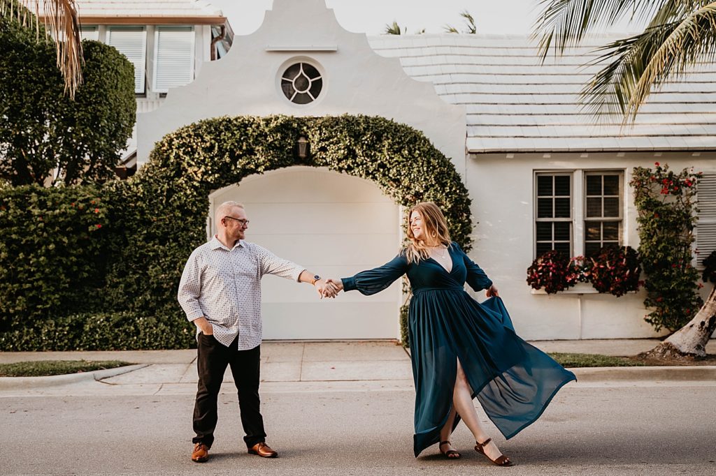 Couple extending their arms out to holding hands as lady curtsies in front of shape greenery hedges and house Worth Ave Palm Beach Engagement Photography captured by South Florida Engagement Photographer Krystal Capone Photography