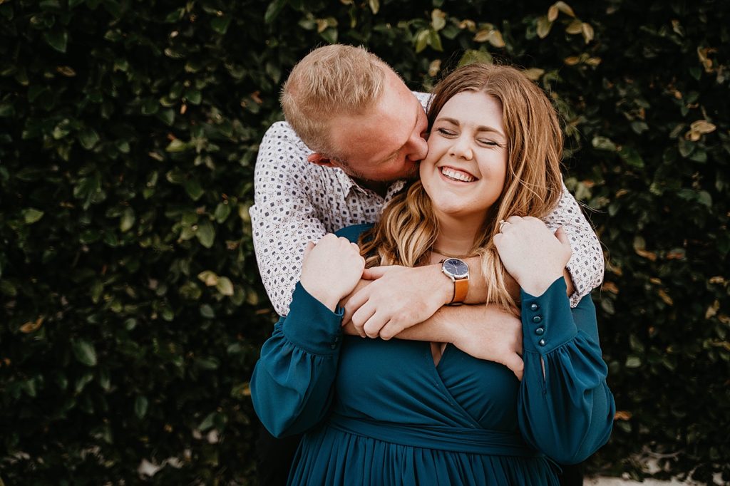 Man hugging and kissing lady from behind in front of hedge Worth Ave Palm Beach Engagement Photography captured by South Florida Engagement Photographer Krystal Capone Photography