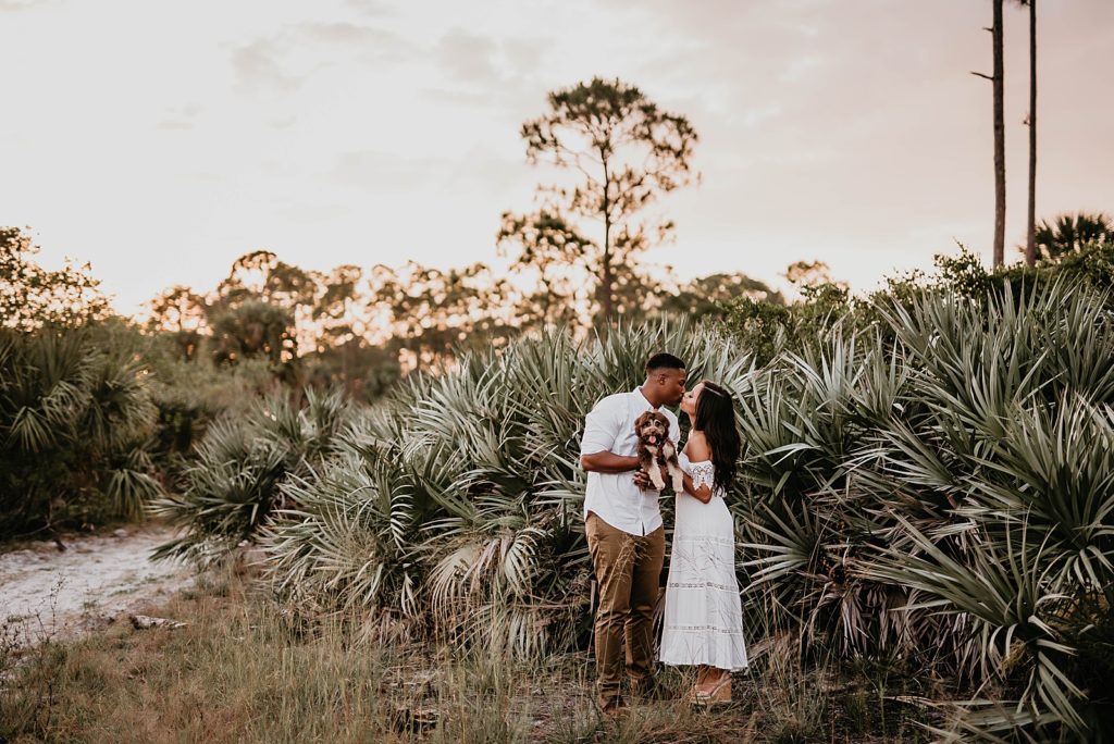 Couple kissing by greenery and holding dog