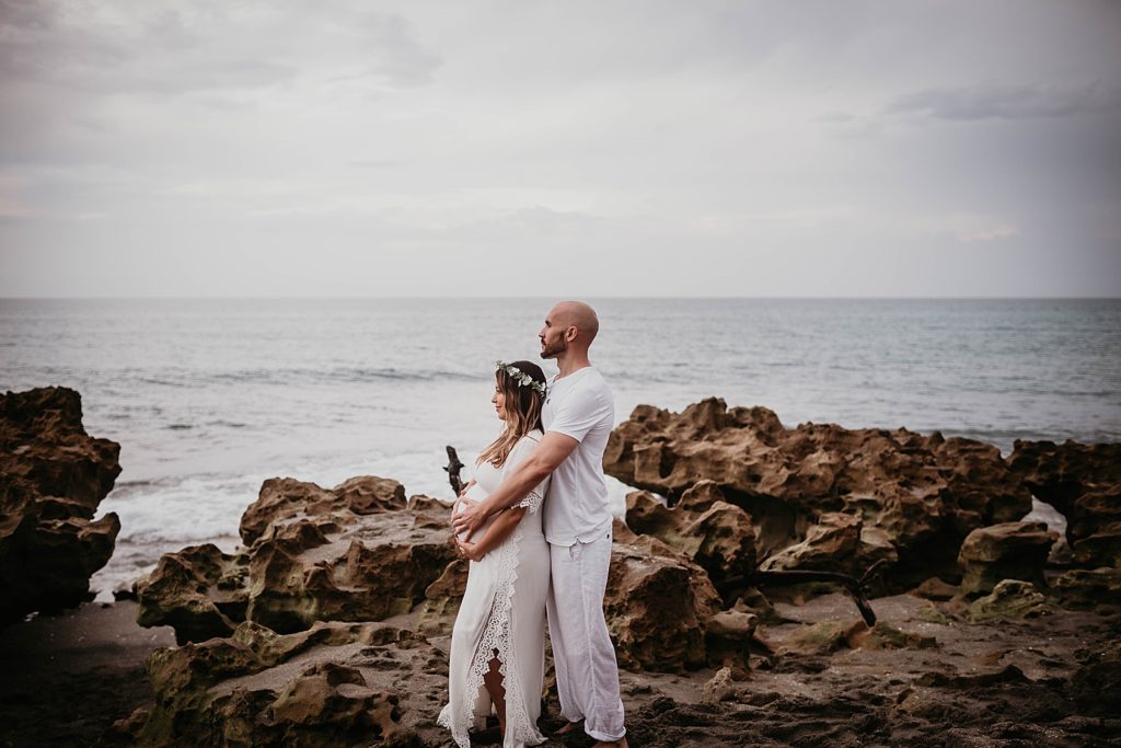 Side profile of Man holding pregnant woman on beach rocks with ocean by them