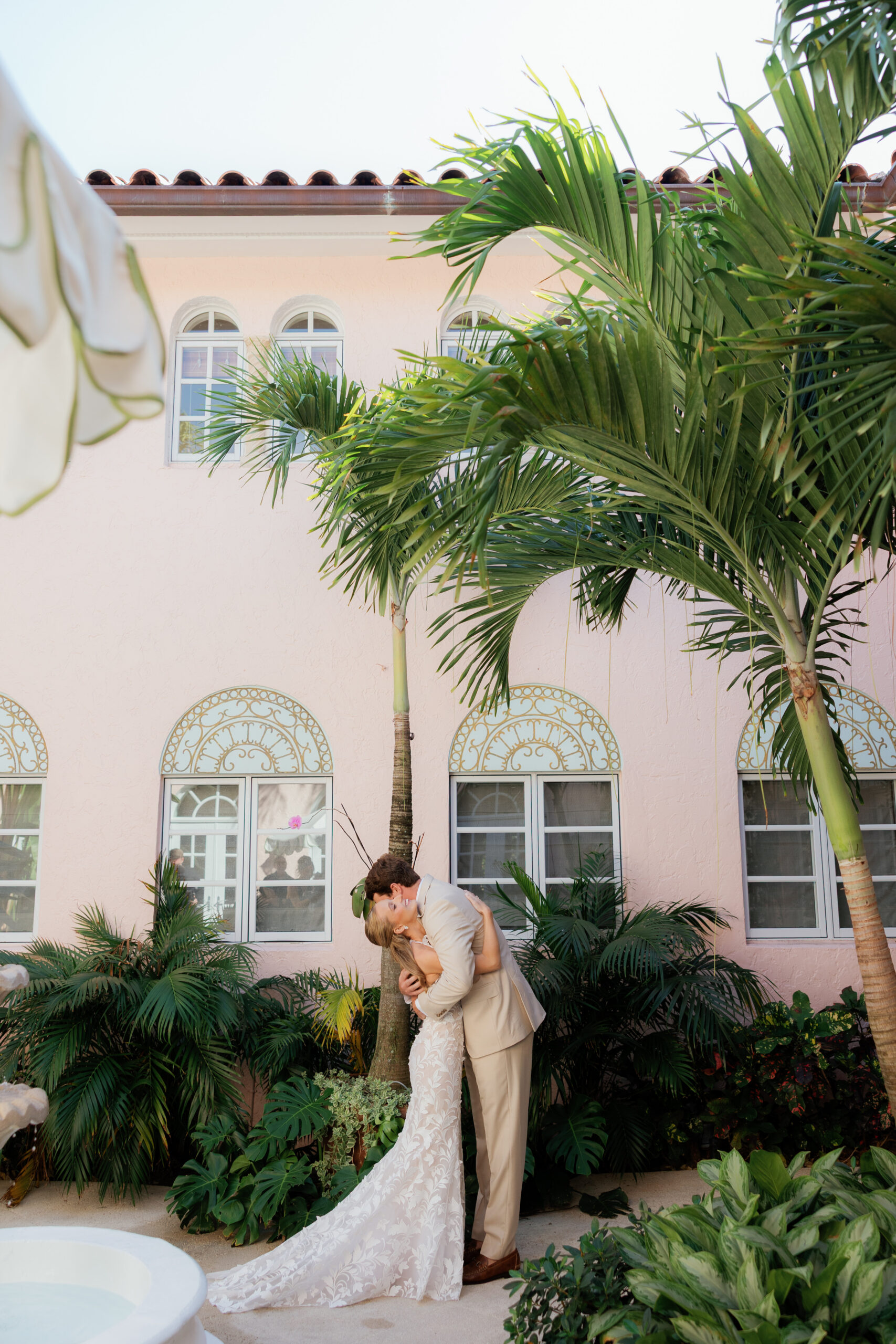 Harper and Luke wedding at The Colony Palm Beach - getting ready - A bride and groom share a kiss outdoors amidst lush greenery and a pink building
