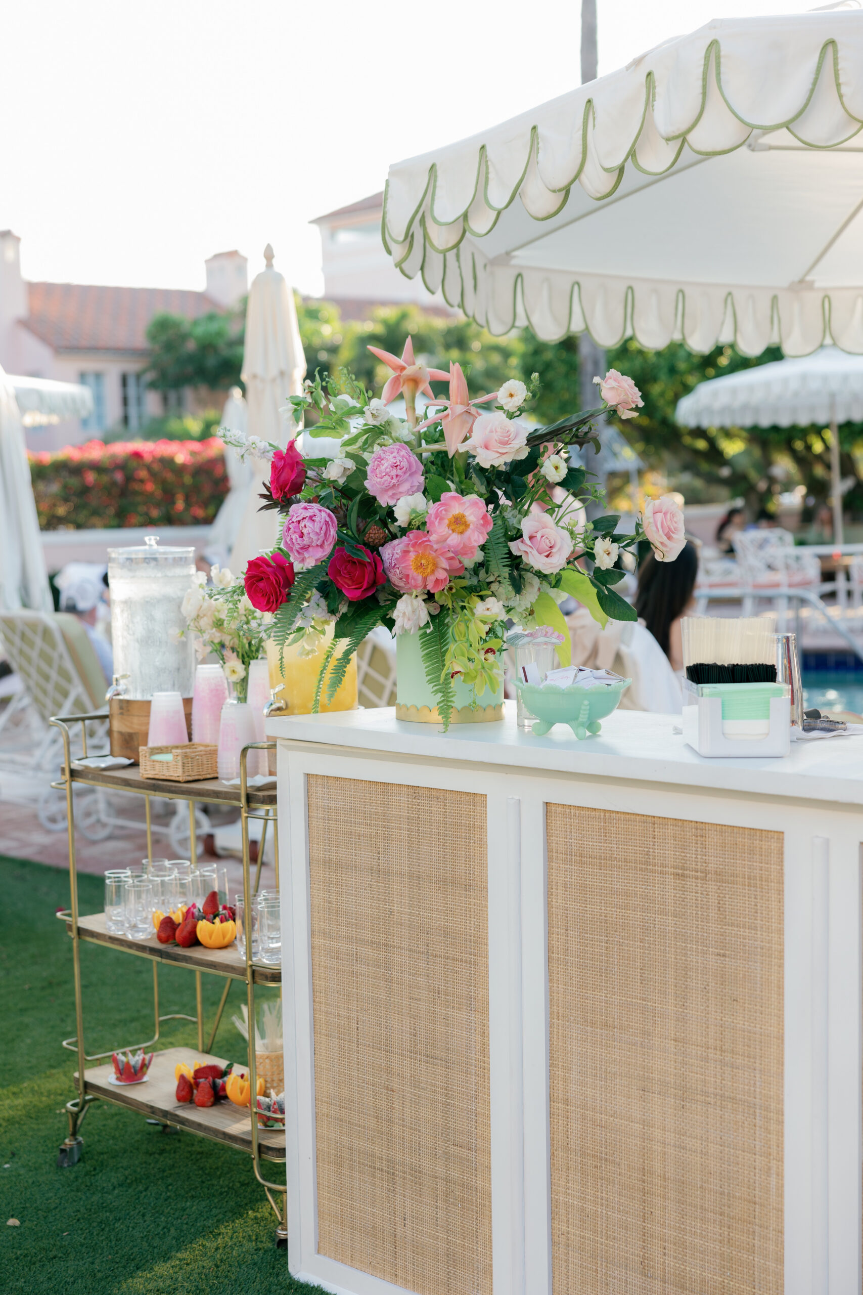Harper and Luke wedding at The Colony Palm Beach - cocktail hour - A bar setup with floral arrangements, drinks, and fruit under an umbrella in an 
