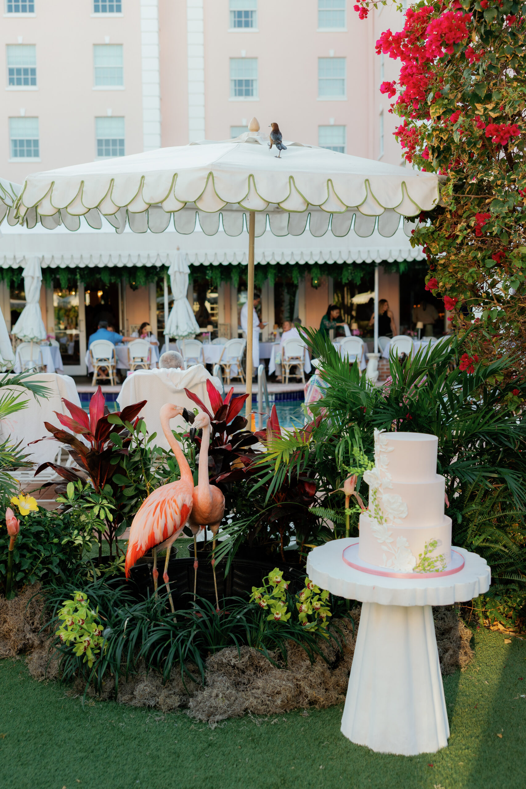 Harper and Luke wedding at The Colony Palm Beach - cocktail hour - A three-tiered wedding cake and flamingo statues are displayed outdoors, with gu