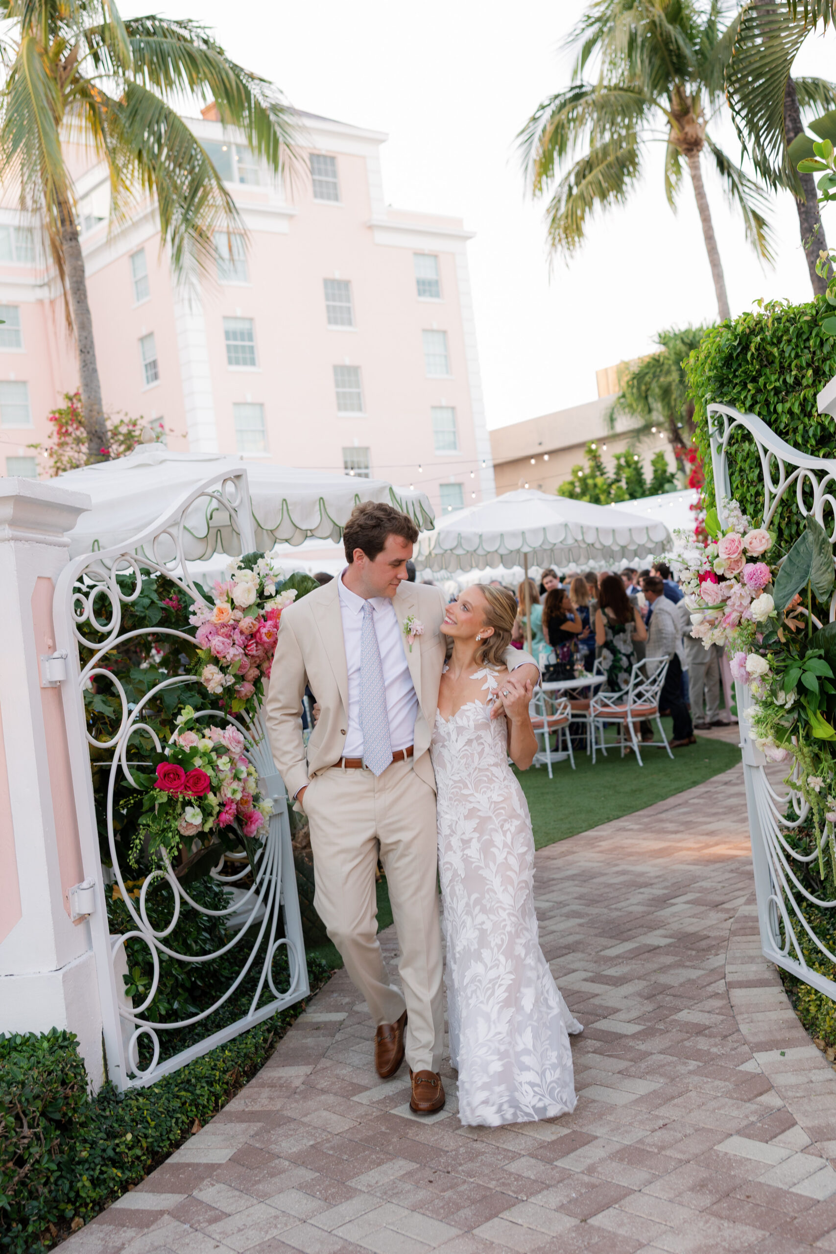 Harper and Luke wedding at The Colony Palm Beach - cocktail hour - A bride and groom walk arm in arm through an outdoor area with guests mingling i