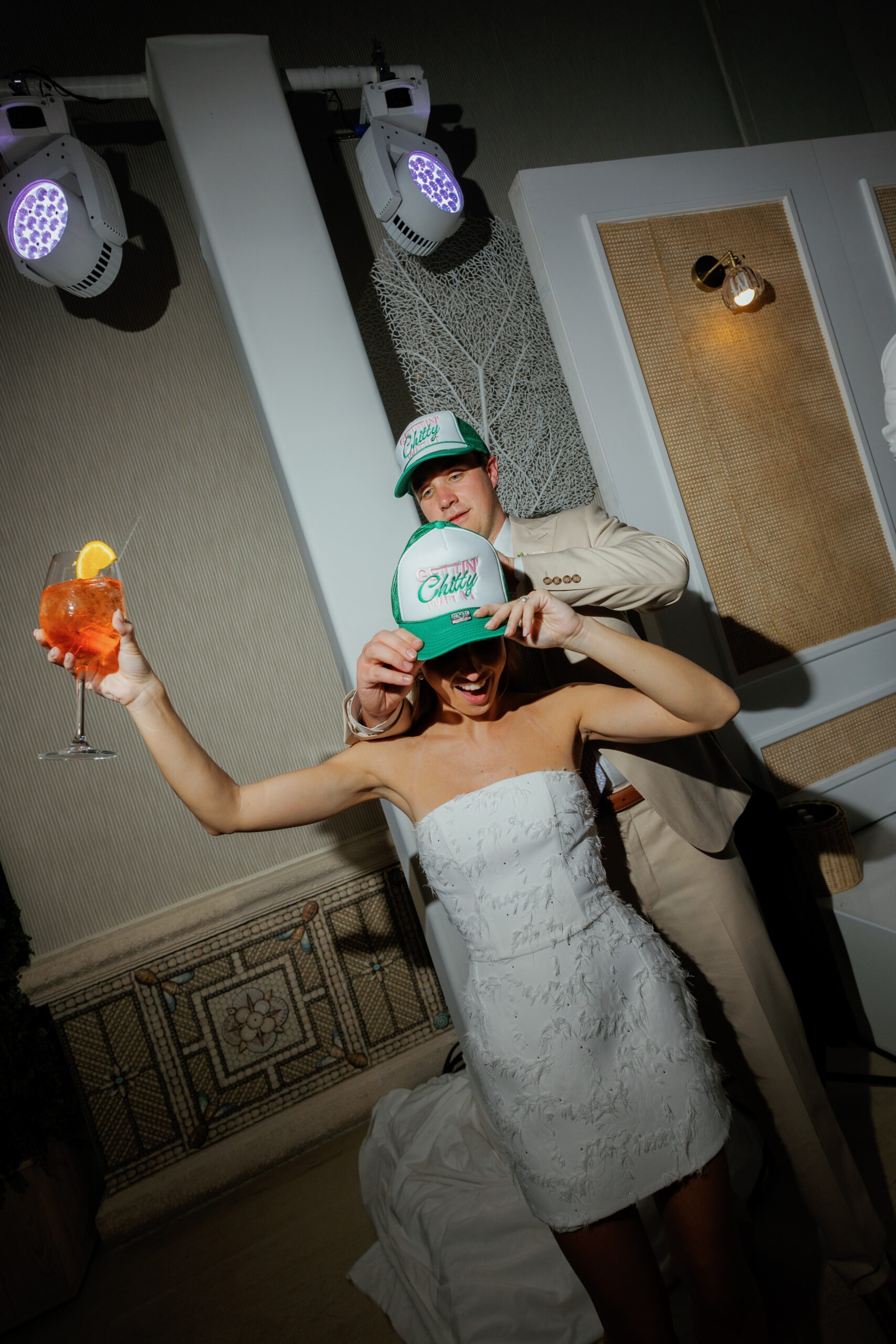 Harper and Luke wedding at The Colony Palm Beach - reception - A couple in party hats and a white dress, holding a drink, under spotlights at a
