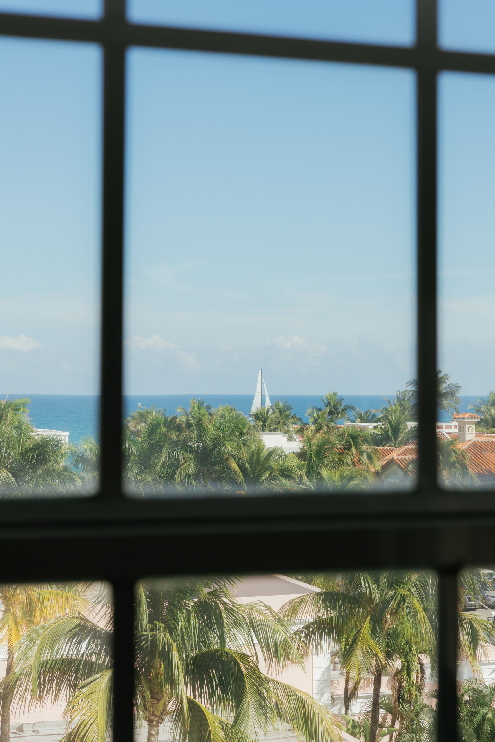 Harper and Luke wedding at The Colony Palm Beach - getting ready - A scenic view of the ocean with a sailboat and palm trees, framed by a window.