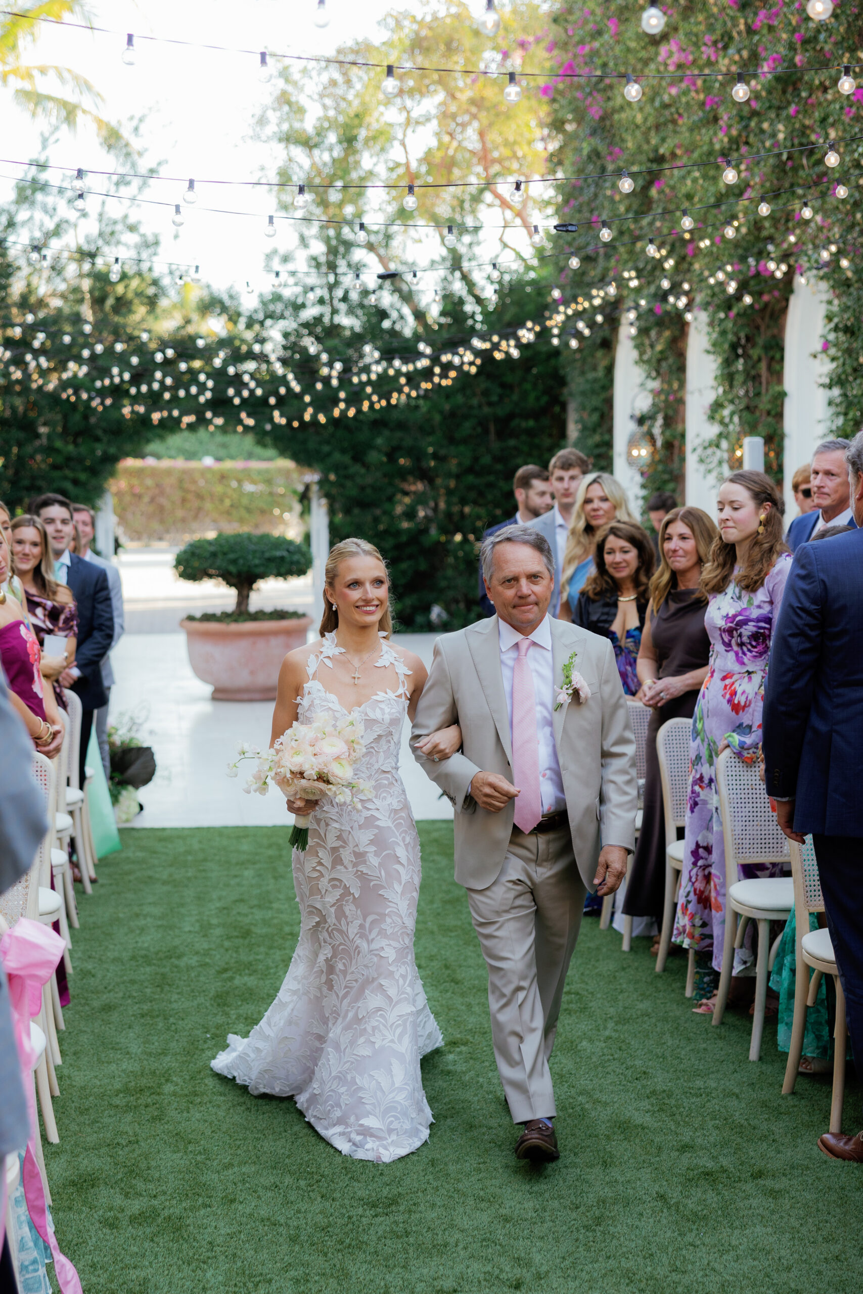Harper and Luke wedding at The Colony Palm Beach - ceremony - A bride in a white dress walks down an aisle with a man, surrounded by seated gu