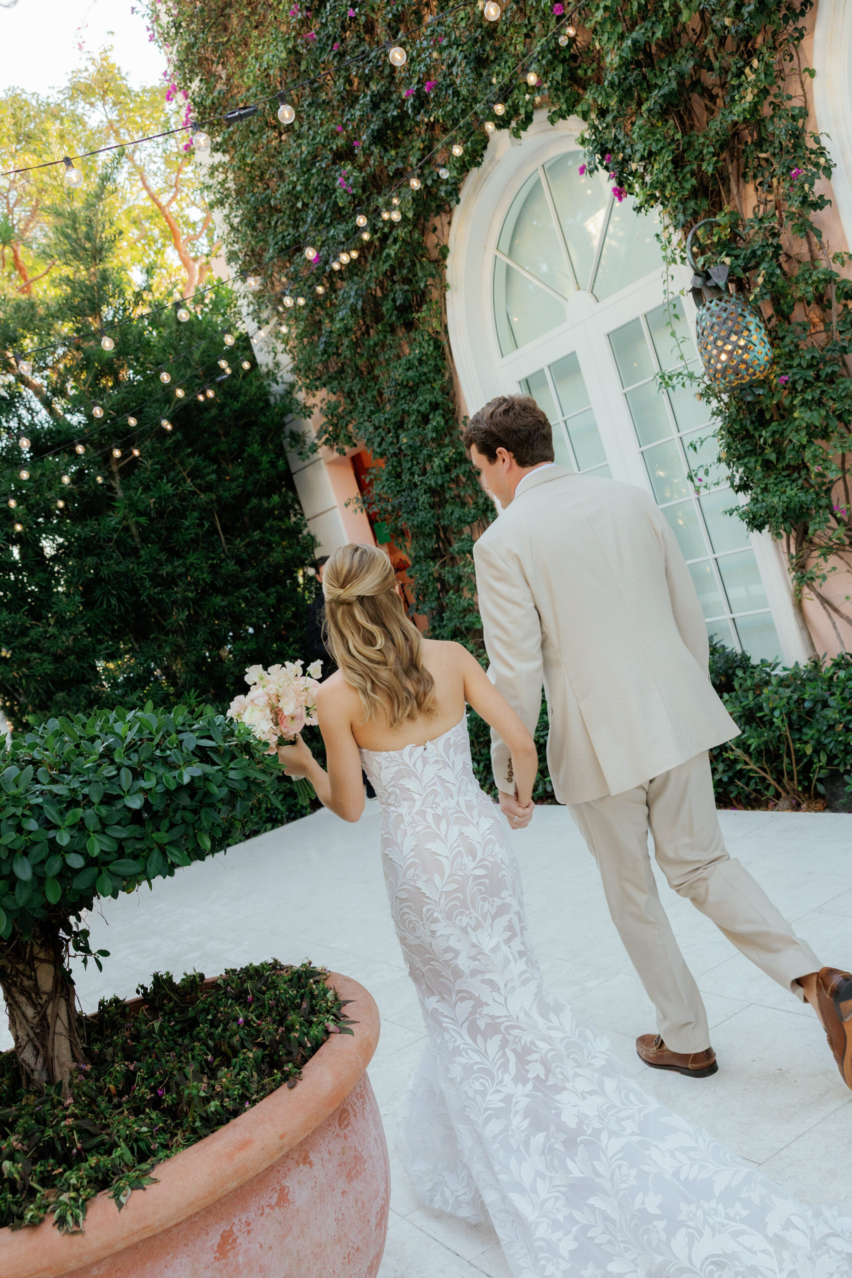 Harper and Luke wedding at The Colony Palm Beach - reception - A bride and groom walk hand-in-hand through an outdoor area adorned with string 
