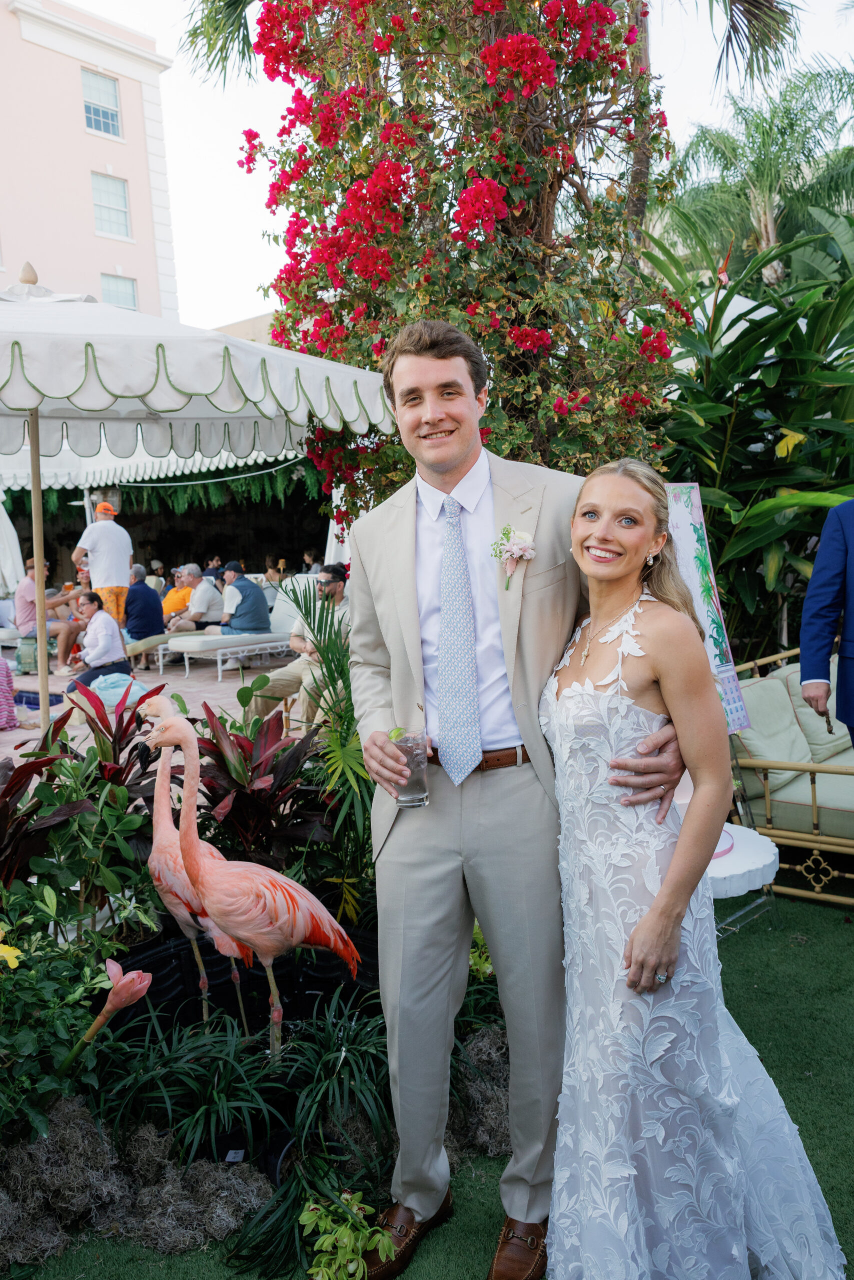 Harper and Luke wedding at The Colony Palm Beach - cocktail hour - A couple poses with flamingos and guests mingling in the background during a coc