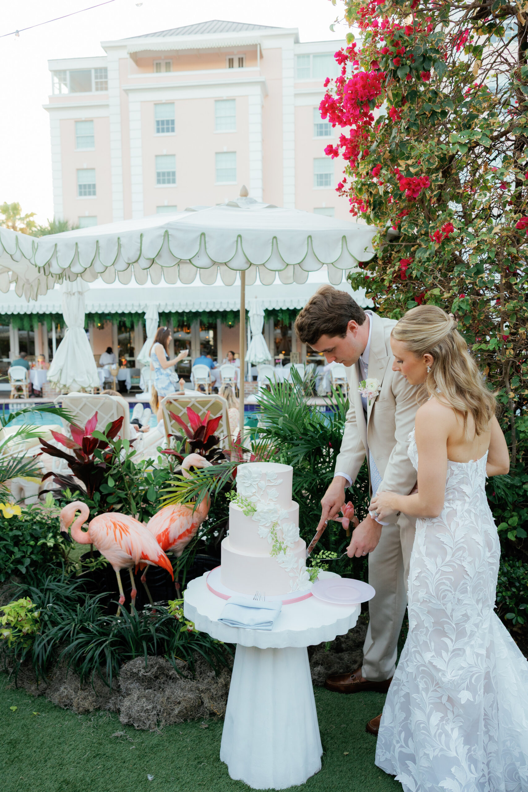 Harper and Luke wedding at The Colony Palm Beach - reception - Bride and groom cut a multi-tiered wedding cake outdoors, with flamingos and gue