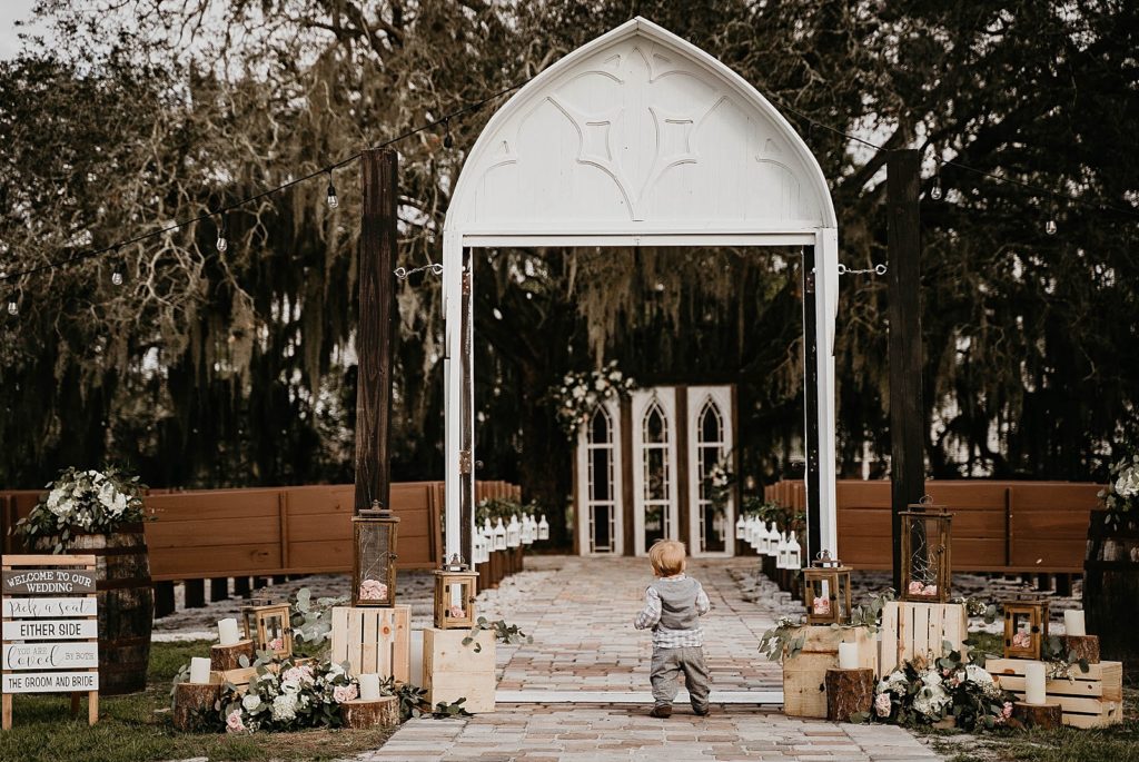 Extravagantly decorated Ceremony area with toddler standing at entrance of aisle Ever After Farms Wedding Photography captured by South Florida Wedding Photographer Krystal Capone Photography 