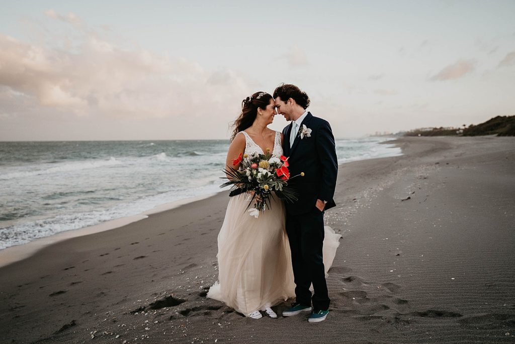 Bride and Groom standing on the beach with tropical bouquet with sneakers on Jupiter Beach Resort Wedding Photography captured by South Florida Wedding Photographer Krystal Capone Photography 