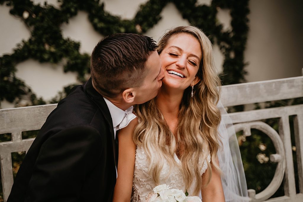 Groom kissing Bride sitting on bench Pelican Club Wedding Photography captured by South Florida Wedding Photographer Krystal Capone Photography