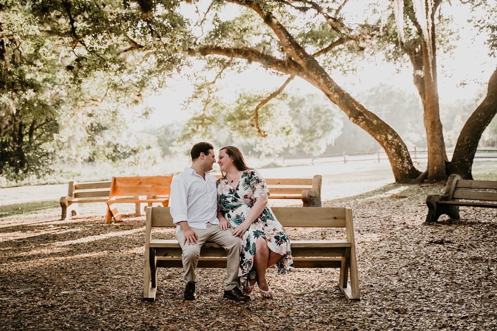 Couple sitting on wooden bench together about to kiss with radiant sun beams behind them Riverbend Park Engagement Photography captured by South Florida Engagement Photographer Krystal Capone Photography 
